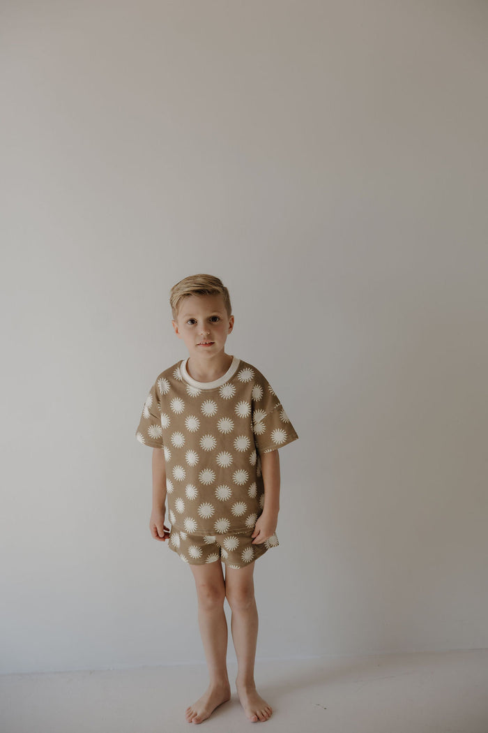 A young boy with short blond hair stands barefoot against a light wall, wearing the French Terry Short Set | Sol by forever french baby. The set features matching shorts and a short sleeve top with a white sunburst pattern.