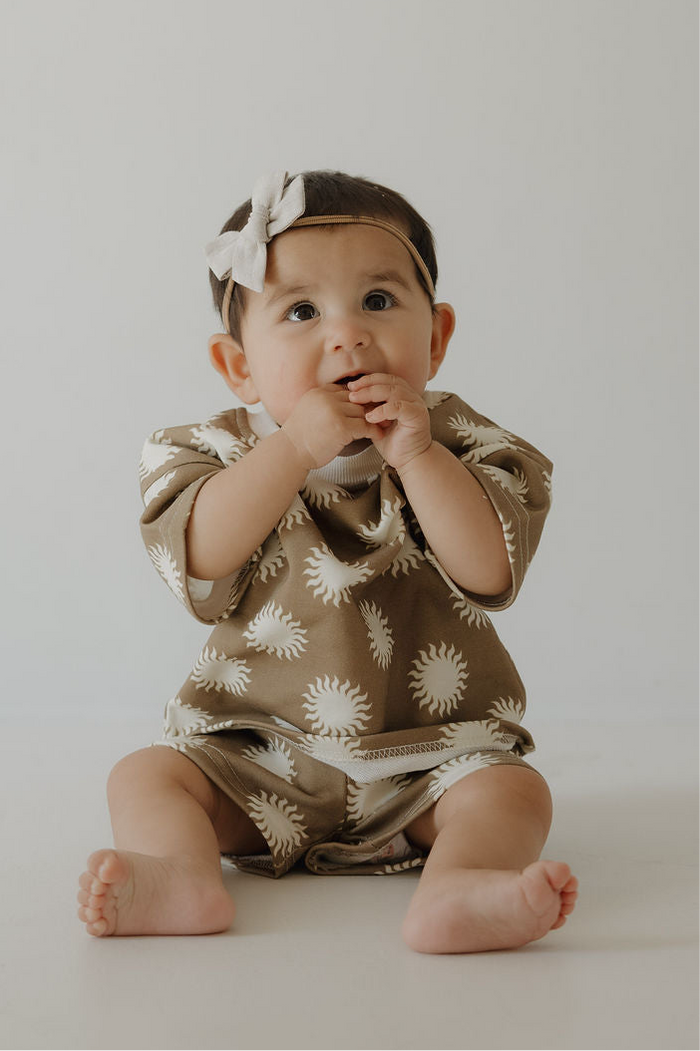 A baby in the forever french baby French Terry Short Set | Sol—with a brown, white sun-patterned top and matching shorts—sits on the floor, hands near mouth, wearing a light bow headband against a plain background.