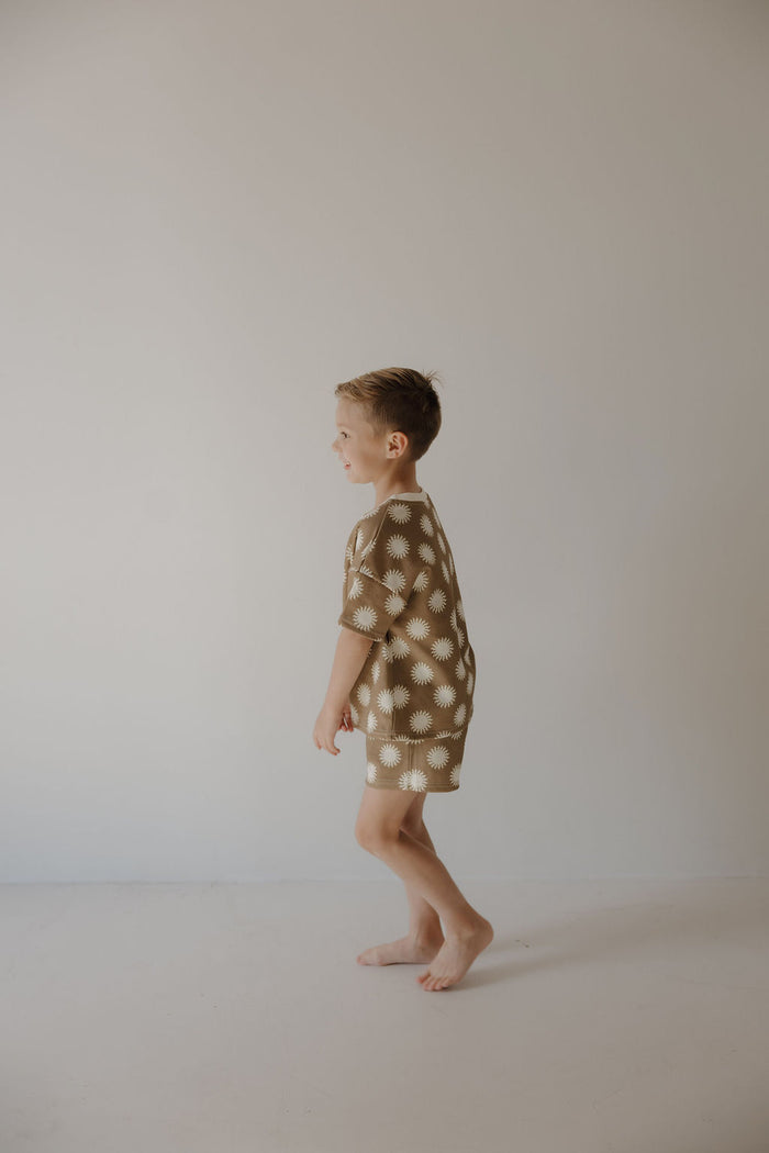 A young boy, barefoot and smiling, stands against a light wall wearing the forever french baby French Terry Short Set | Sol—brown short sleeves and matching shorts with white daisy patterns—looking to his right.