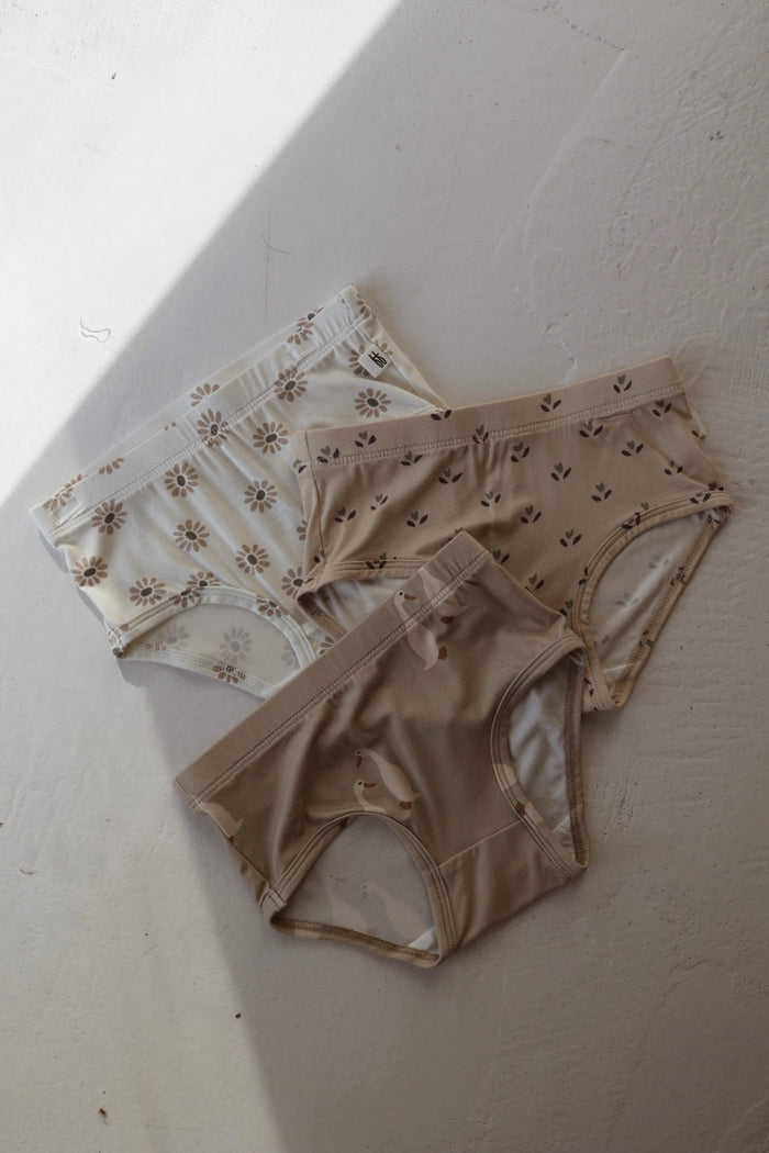 Three pairs of forever french baby Girl’s Bamboo Briefs (3 Pack) in Flora, Silly Goose & Daisy Fields prints are displayed on a light, textured surface with partial shadow. Hypo-allergenic and in neutral tones with fun patterns.
