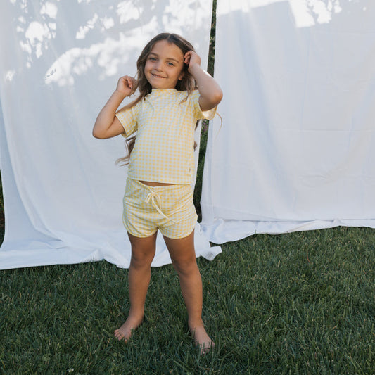 A young girl stands barefoot on grass, smiling and holding her long hair. She wears the forever french baby Girl's Bamboo Short Set in Citron, with a white fabric backdrop outdoors.