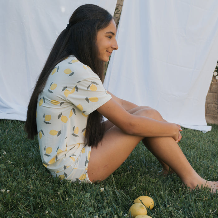 A person with long dark hair sits cross-legged on grass, wearing the forever french baby Girl's Bamboo Short Set | Lemon. Hypo-allergenic material adds comfort, with lemons nearby and white fabric in the background.