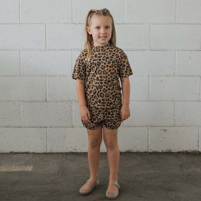 A young girl stands in front of a white brick wall, wearing the forever french baby Girl's Bamboo Short Set | Leopard—breathable bamboo T-shirt and shorts—with clear plastic shoes. She faces forward and smiles slightly.