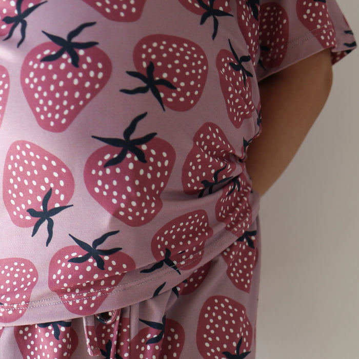 Close-up of a person wearing the Girl's Bamboo Short Set | Matilda by forever french baby, featuring a pink and purple strawberry pattern, showing their torso and arm with hand tucked behind their back.