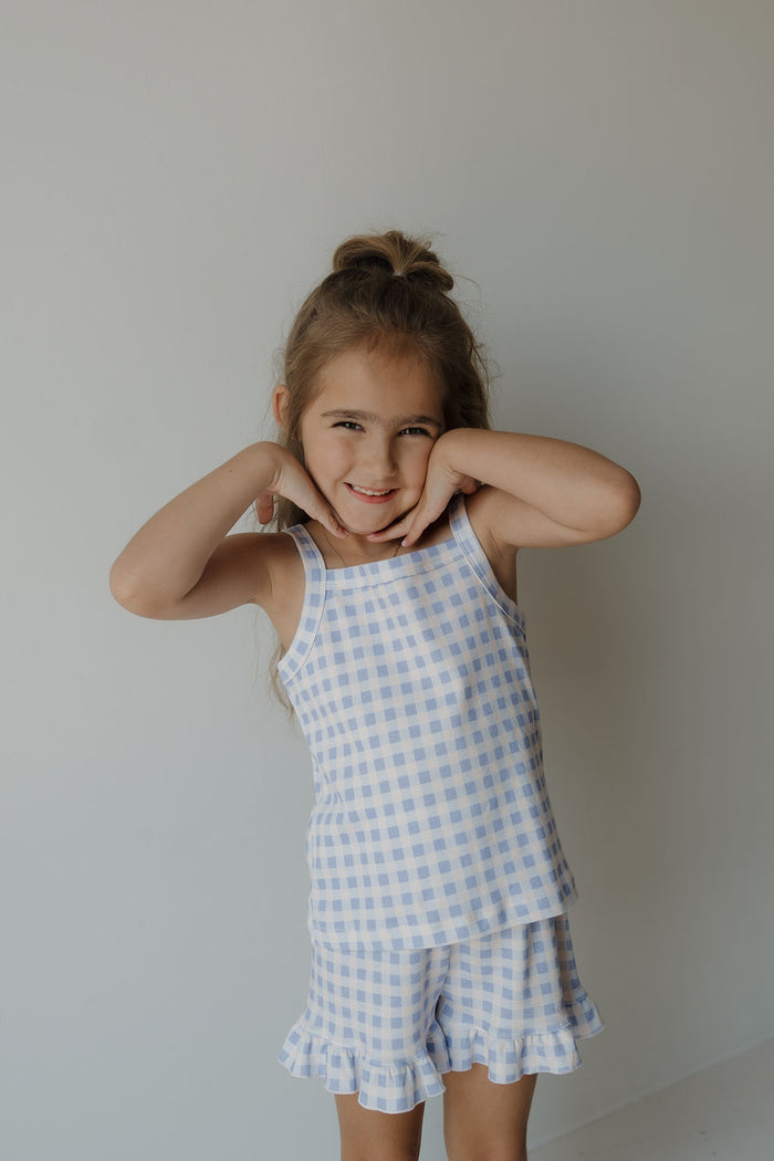 A young girl with long hair styled half-up poses playfully, smiling and touching her face. She wears the forever french baby Girl’s Ruffle Short Set | Marilyn in a blue and white gingham print cotton blend, standing against a plain, light-colored background.