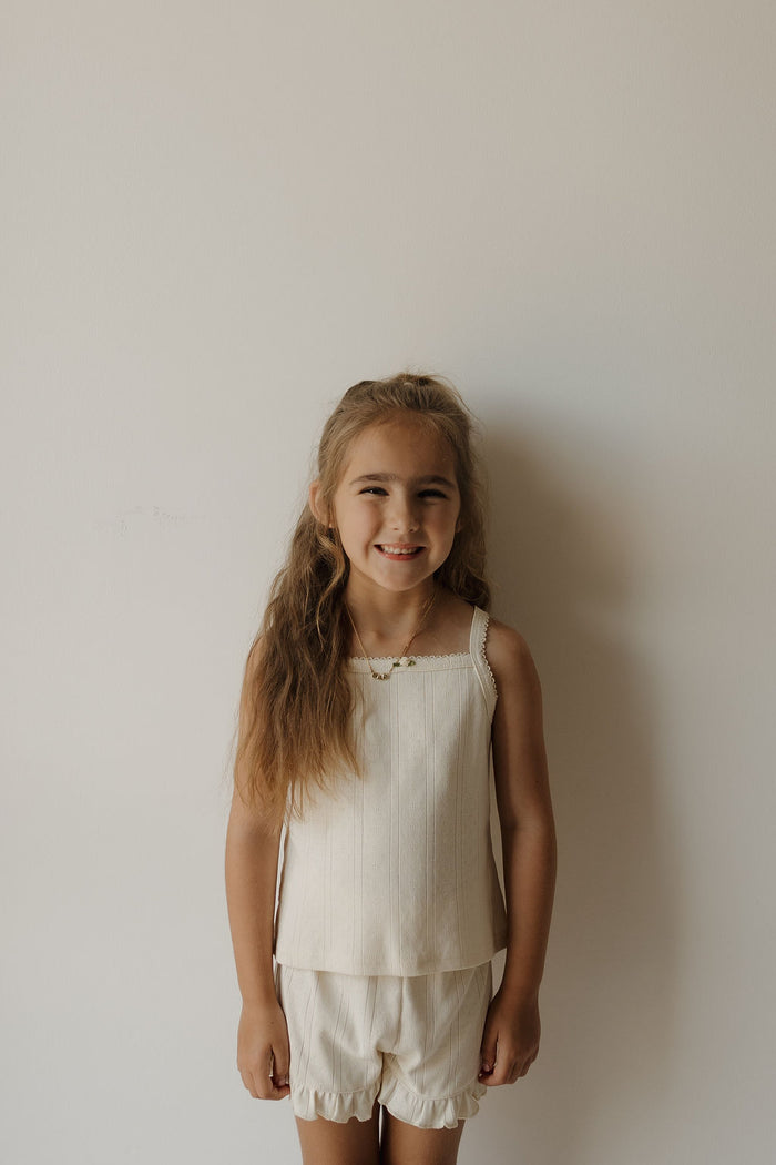 A young girl with long light brown hair stands against a plain white wall, smiling softly as she wears the forever french baby Girl’s Ruffle Short Set | Swan—a sleeveless cream top and matching ruffle shorts made from soft pointelle fabric.