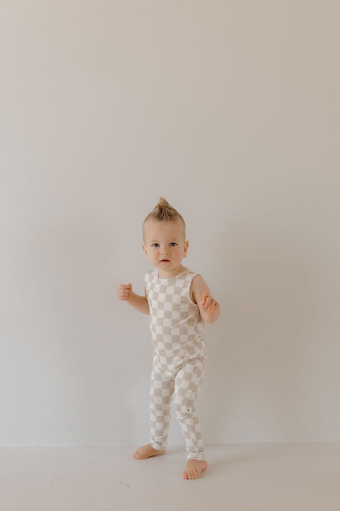 A baby stands barefoot against a neutral background in a sleeveless Harem Bamboo Romper by forever french baby, showcasing a beige and white checkered design. With a small tuft of styled hair, the baby gazes curiously at the camera.