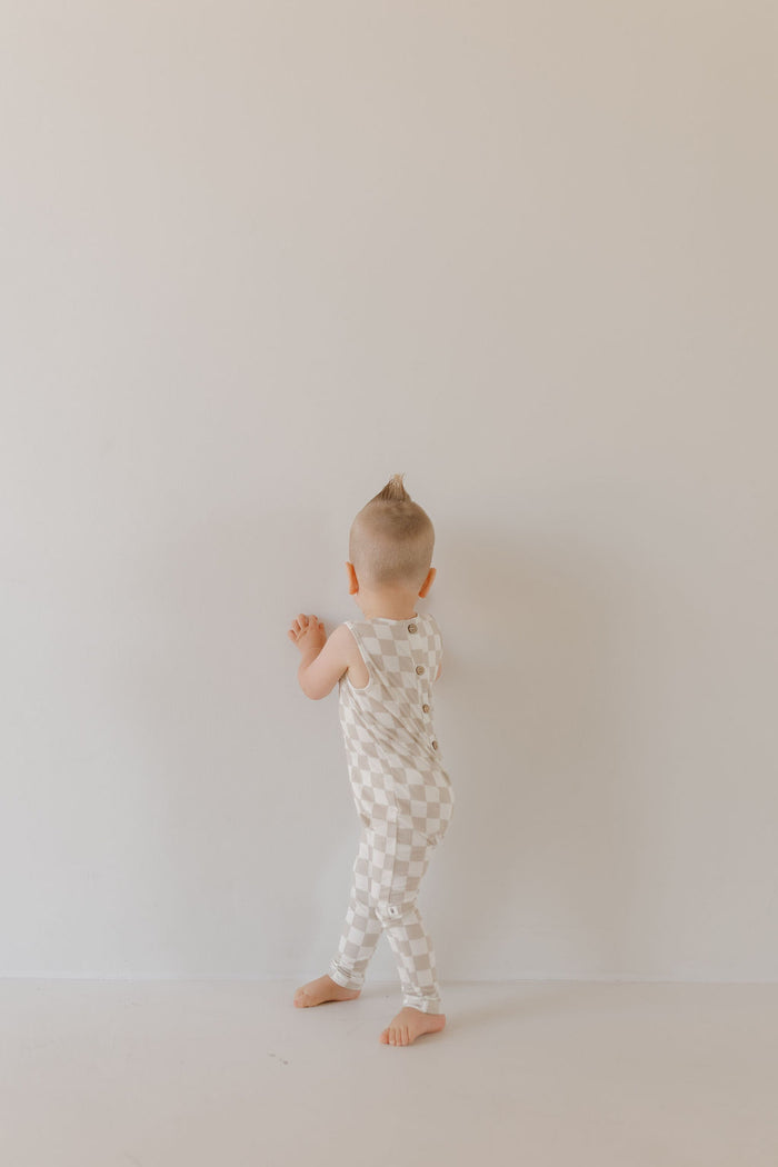 A toddler with a small mohawk wears the Harem Bamboo Romper | Checkerboard by forever french baby, standing barefoot against a plain off-white wall in a minimally decorated room, highlighting the child's playful pose.