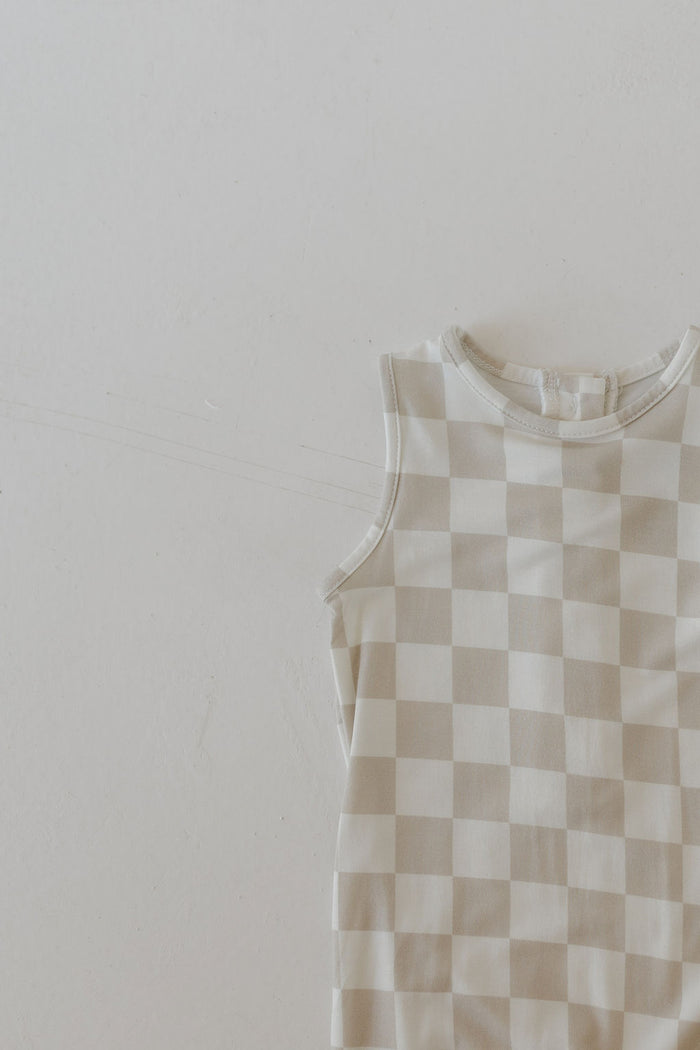 The Harem Bamboo Romper in a beige and white checkerboard pattern by forever french baby is showcased against a plain white background. Crafted from breathable bamboo, this sleeveless romper is neatly folded to highlight its simple and minimalistic design.