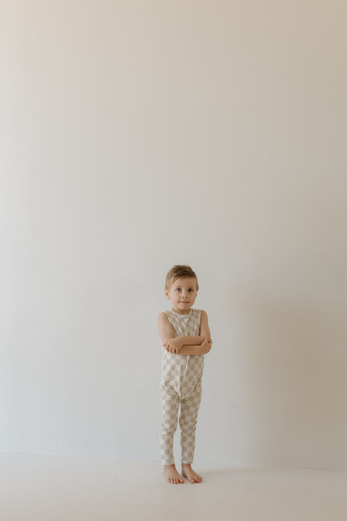 A young child with short hair and a neutral expression stands barefoot with crossed arms, wearing the Harem Bamboo Romper | Checkerboard by forever french baby. The setting is minimalistic against an off-white wall.
