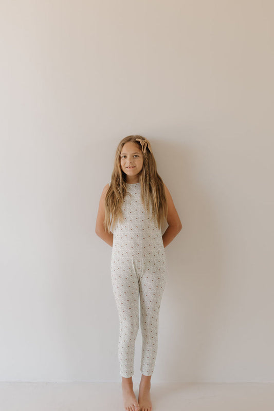 A girl with long hair stands barefoot against a plain white wall, wearing the Harem Bamboo Romper in Coast by forever french baby. With a slight smile and hands behind her back, she complements the minimalist, neutral background, and the romper's breathable fabric enhances the serene simplicity.