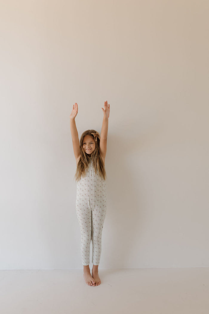 A young girl in a white Harem Bamboo Romper by Forever French Baby stands barefoot against a plain wall, arms raised and smiling.