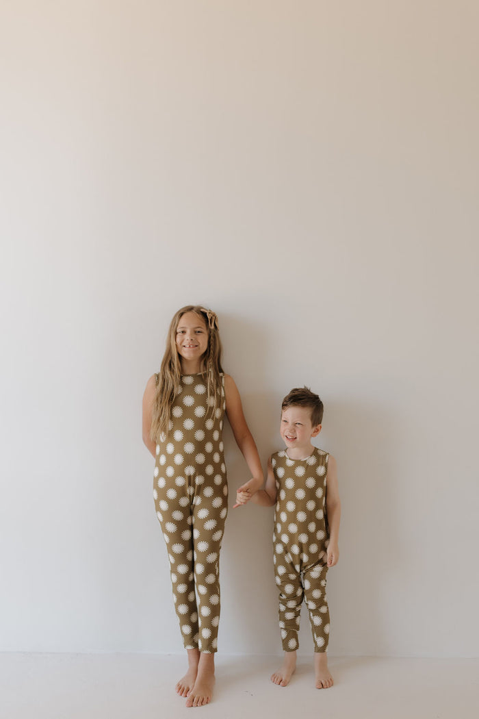 Two children hold hands against a plain backdrop, wearing matching "Harem Bamboo Romper | Sol" outfits by forever french baby. The brown rompers with white polka dots are breathable and hypo-allergenic for comfort. One child has long hair; the other has short locks. Both smile barefoot.