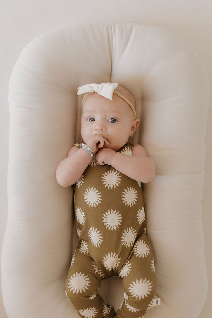 A baby in the forever french baby Harem Bamboo Romper | Sol lies on a soft, beige cushion. The brown onesie with white sun patterns, made from breathable bamboo fabric, ensures comfort and care. A bow-adorned headband completes the look against a minimal, soft-toned background.