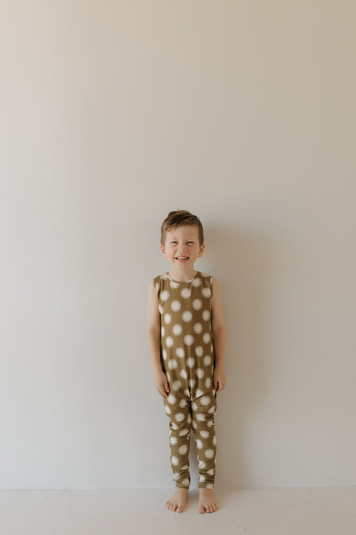 A young child stands barefoot against a plain, light background, smiling in a sleeveless Harem Bamboo Romper | Sol by forever french baby. This olive-green romper with white flowers is hypoallergenic and perfect for sensitive skin, offering an adorable blend of style and comfort.