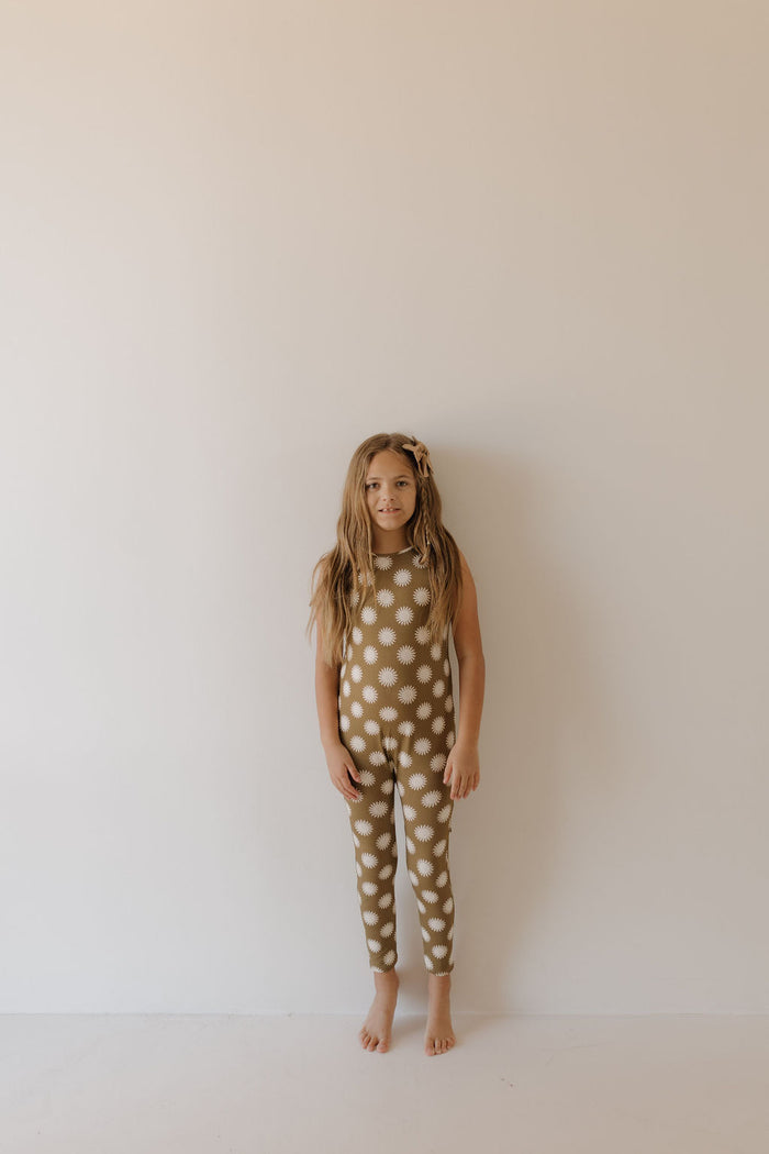 A young girl poses barefoot before a plain off-white wall, wearing the charming Harem Bamboo Romper in Sol from forever french baby. Her loose hair and focused gaze highlight the sleeveless and stylish design, crafted from breathable bamboo for ultimate comfort.
