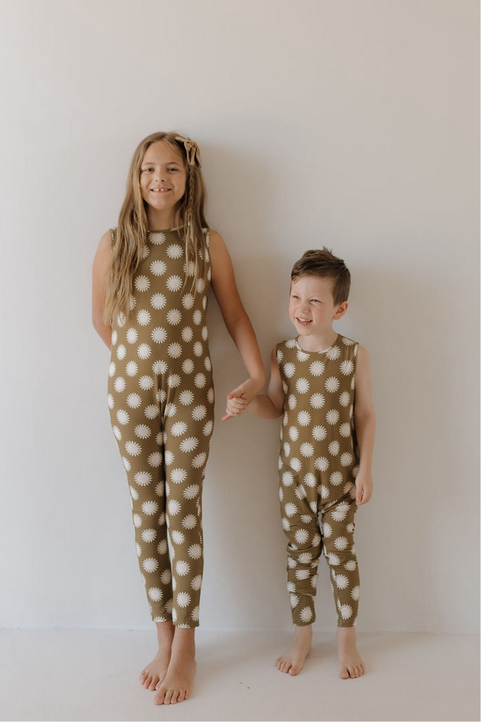 Two young children stand barefoot against a white wall, holding hands and smiling in matching forever french baby Harem Bamboo Rompers | Sol—daisy print outfits designed for comfort, playfulness, and breathable style.