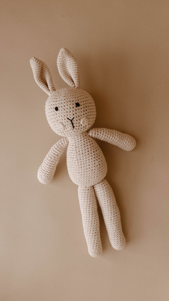 A beige knit bunny doll named Honey the Bunny from forever french baby lies on a light brown surface. The bunny features long ears, a simple stitched face with two eyes, a small nose, and a slight smile. Its elongated arms and legs give it a cuddly, handmade appearance.