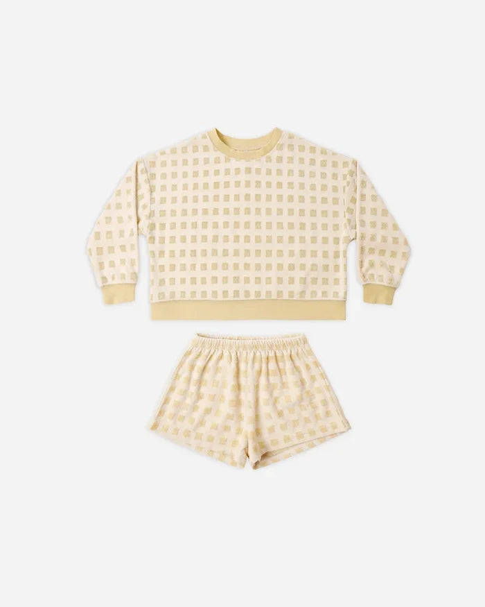 Introducing the Karli Set by Rylee + Cru: a coordinated ensemble featuring a cream-colored, long-sleeve sweatshirt and matching shorts adorned with a subtle light yellow checkered pattern. Crafted from soft terry cloth, this set offers an easygoing coastal vibe and is displayed laid flat on a white background.