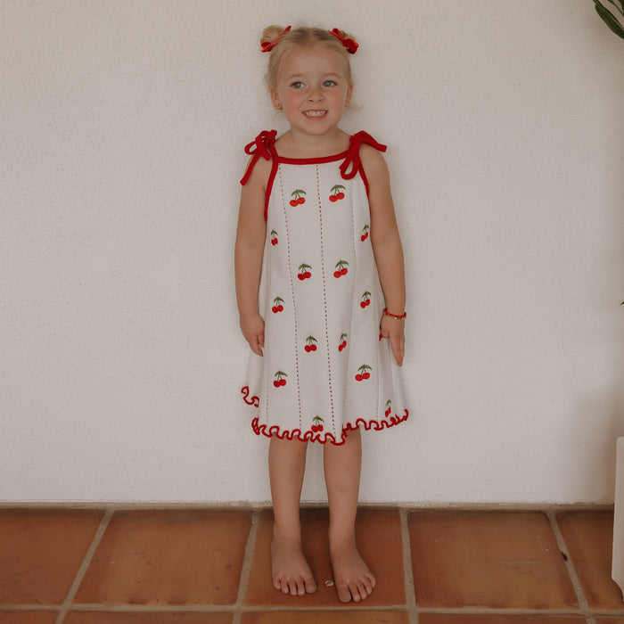 A young girl with blonde pigtails and red bows stands barefoot on terracotta tiles, smiling against a white wall. She wears the forever french baby Knit Dress | Cherry, a white 100% cotton dress with red trim and cherry prints.