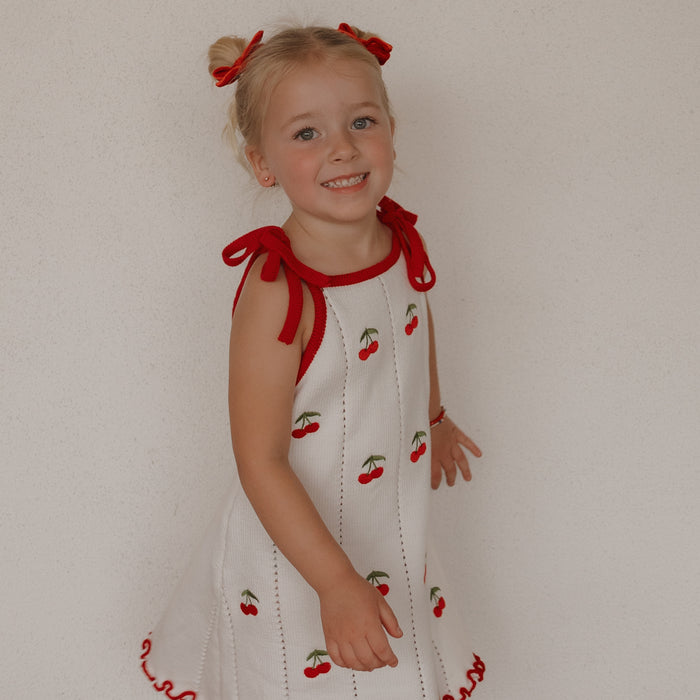 A smiling young girl with blonde pigtails tied with red bows wears the forever french baby Knit Dress | Cherry, a cute 100% cotton white dress with cherry patterns and red trim, standing before a plain light-colored wall.