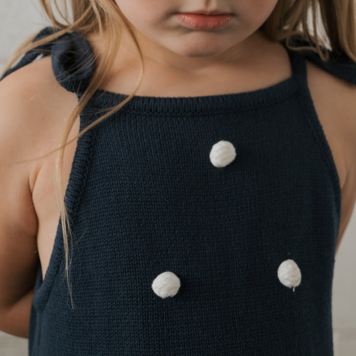A young child wears the forever french baby Knit Dress | Emery—navy blue, 100% cotton, sleeveless with adjustable tie straps and three white pom-poms—standing against a neutral background.