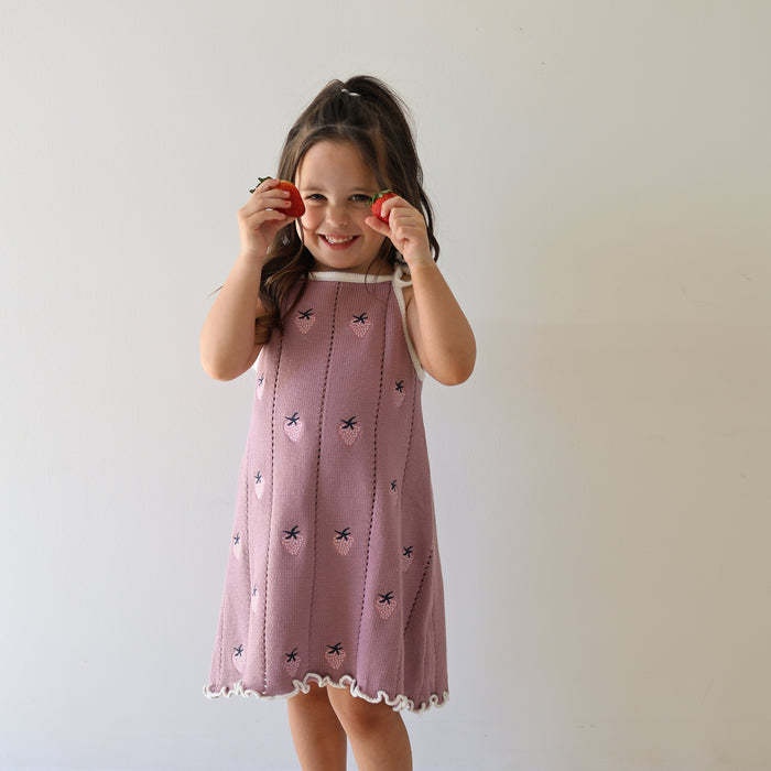 A young girl smiles while holding two strawberries to her eyes, wearing the forever french baby Knit Dress | Matilda—a pink 100% cotton knit dress with a scalloped hem and adjustable tie straps—against a plain white background.