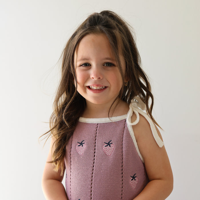 A young girl with long brown hair wears the forever french baby “Knit Dress | Matilda,” a purple sleeveless dress with adjustable tie straps, and smiles at the camera against a plain white background.