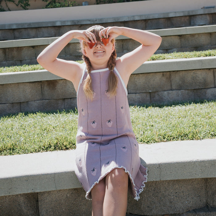 A young girl wears the forever french baby Knit Dress | Matilda with adjustable tie straps as she sits on outdoor steps, playfully holding peach slices over her eyes in the sunlight, with grass and a stone wall in the background.