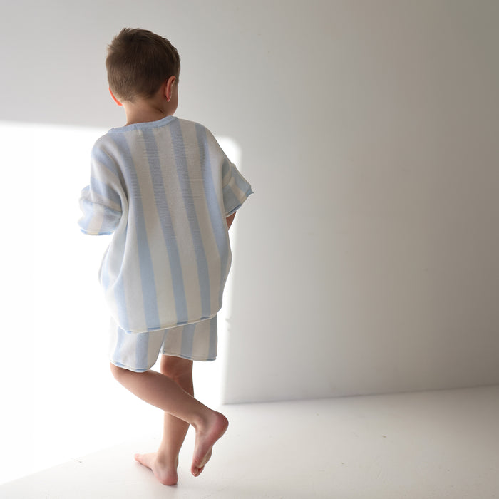 A young child stands barefoot in soft sunlight, facing away, wearing the forever french baby Knit Short Set | Benny—100% cotton with light blue and white cabana stripes—in a minimalist white room.