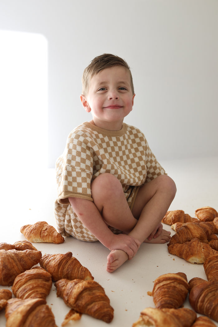 A young child in a forever french baby Knit Short Set | Latte Check sits barefoot on the floor, smiling and surrounded by croissants in a bright, minimalistic room.