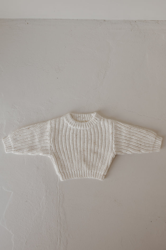 The Child Knit Sweater | Cloud Confetti by forever french baby is a cream-colored, hand-knitted sweater with long sleeves, a ribbed pattern, and a round neckline, shown laid flat on a textured light gray surface.