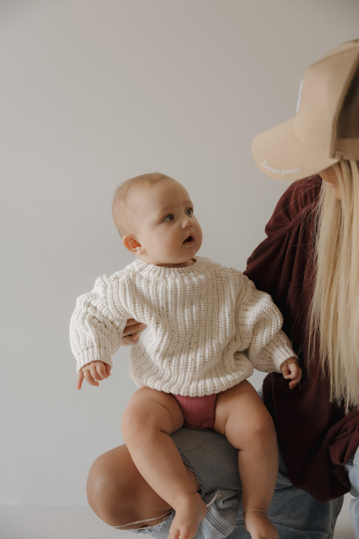 A baby in the forever french baby Child Knit Sweater | Cloud Confetti sits on an adult's lap, featuring a light background.