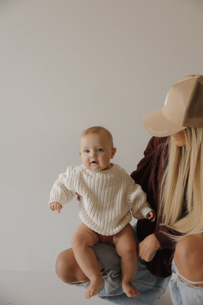 A baby in the forever french baby Child Knit Sweater | Cloud Confetti sits on a person's lap. The person, with long blonde hair and a tan baseball cap, wears casual clothes. The background is light and plain.
