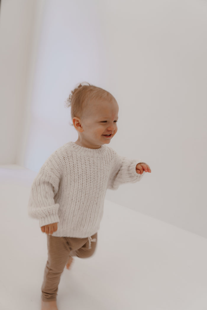 A toddler with a happy expression and short hair walks indoors, wearing the 'Cloud Confetti' knit sweater by forever french baby and brown pants. The softly lit background is minimalistic.