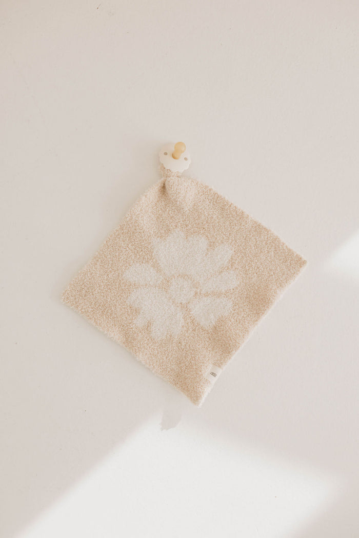 The Lazy Daisy | Lovey by forever french baby is a beige textured microfiber baby lovey with a white flower design, lying flat with a small cream and tan pacifier attached to one corner of the soft fabric.