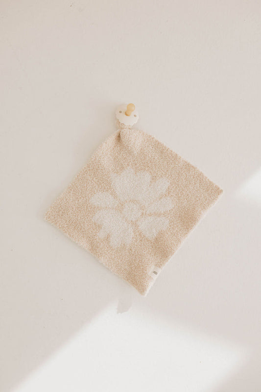 The Lazy Daisy | Lovey by forever french baby is a beige textured microfiber baby lovey with a white flower design, lying flat with a small cream and tan pacifier attached to one corner of the soft fabric.