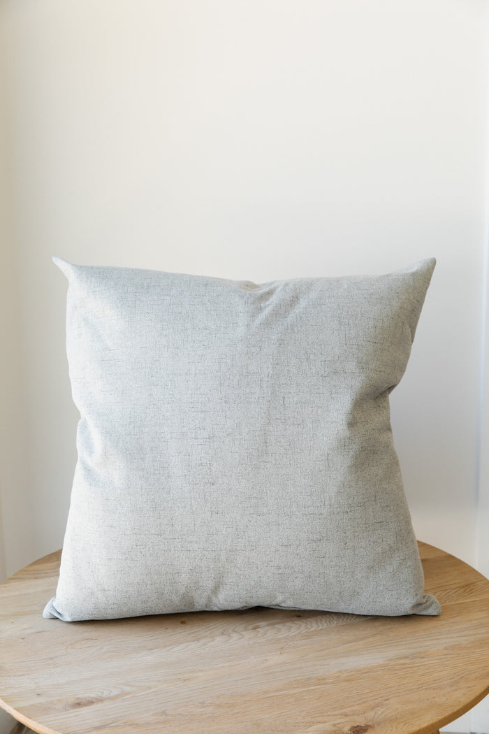 The Limestone | French Pillow, a light grey square cushion with a textured fabric cover and a feather down insert from forever french home, sits on a round wooden table against a plain white wall.