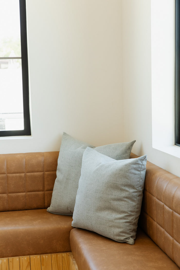 A cozy corner features a brown leather tufted sofa adorned with two large Limestone | French Pillows, boasting feather down inserts. The sofa is situated next to two large windows, allowing natural light to fill the bright and minimalist room, embodying the elegance of Forever French Home.