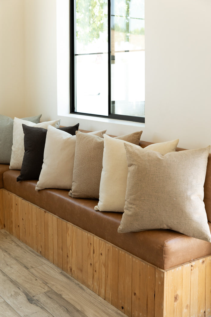 A long wooden bench lined with neutral-colored cushions in shades of beige, brown, and grey sits against a white wall beneath a window. Limestone pillows with feather down inserts from Forever French Home enhance the comfort, while the light-colored wooden floor contributes to a minimalist and cozy atmosphere.