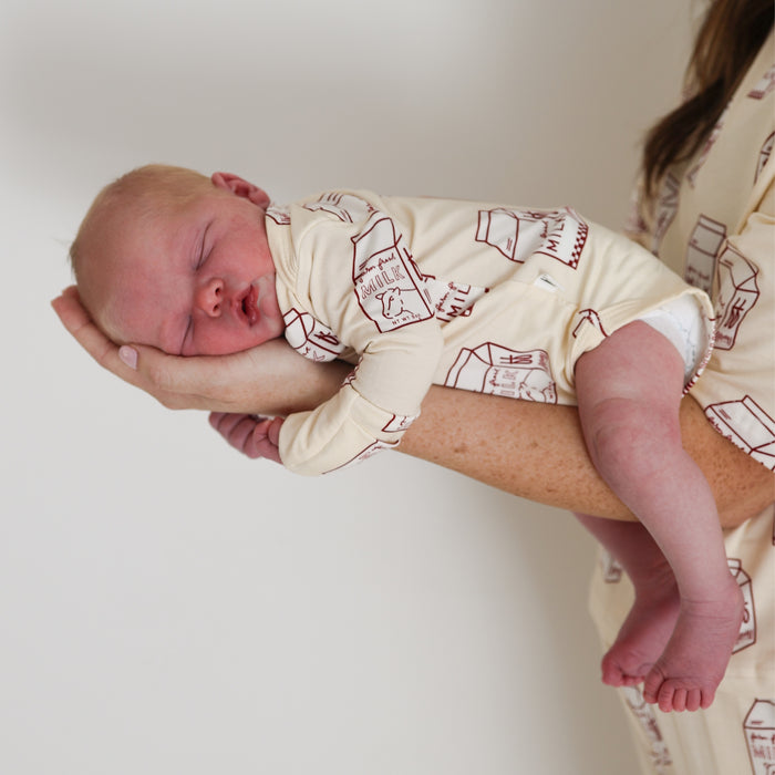 A newborn in a forever french baby Long Sleeve Bamboo Snapsuit | Milk sleeps, cradled gently in an adult's hand and arm, against a plain white background.