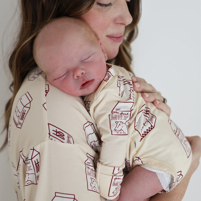 A woman holds a sleeping baby on her shoulder. Both wear matching forever french baby Long Sleeve Bamboo Snapsuits in cream with red milk carton prints. The clothing is hypoallergenic, and the baby's resting peacefully while the woman's face is partly visible.