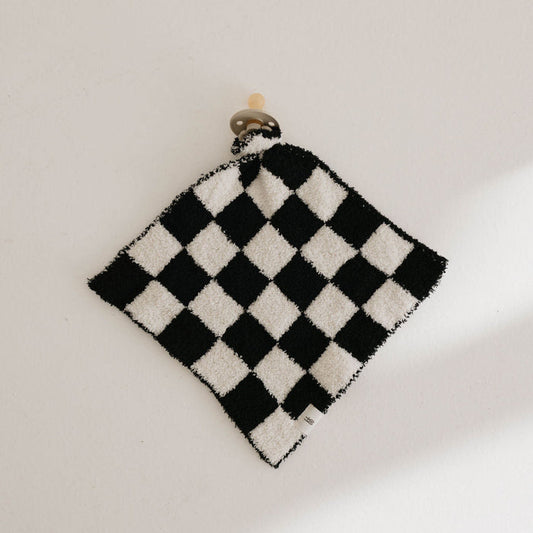 A small Lovey in the Black & White Checkerboard pattern by forever french baby is pinned up against a light-colored wall, secured at the top with a small wooden peg. The microfiber feather yarn gives it a fluffy texture, and there's a visible tag attached to it.