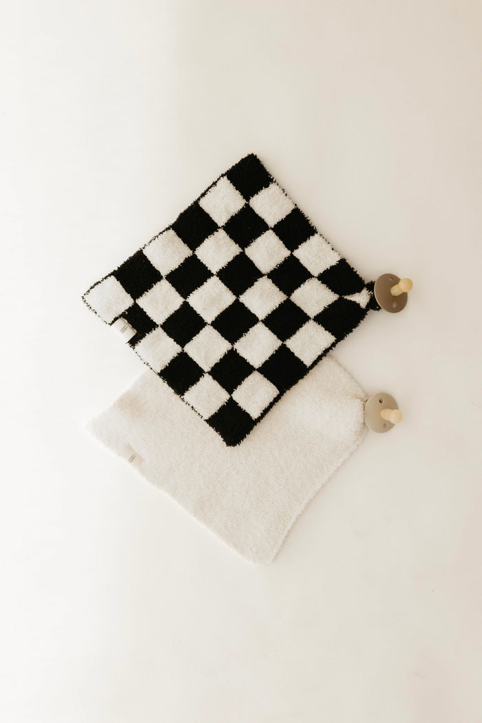 A Lovey by forever french baby, featuring a black and white checkerboard pattern, and a plain white potholder are laid on a light surface. Each is crafted with microfiber feather yarn and features a wooden hook at the corner for hanging.