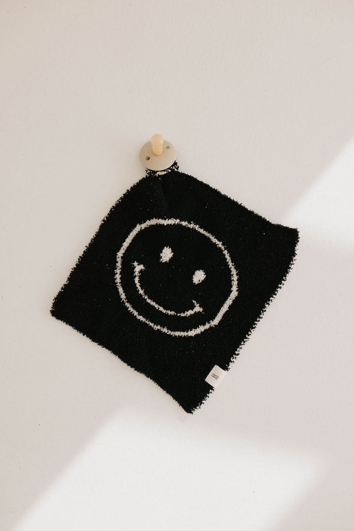 A black square rug made from microfiber feather yarn, featuring a white smiley face design, hangs on a white wall. This rug, named Lovey | Black & White Just Smile by Forever French Baby, is secured by a small wooden peg at the top corner. A small white tag is visible on the bottom edge, and shadows create a pattern on the wall.