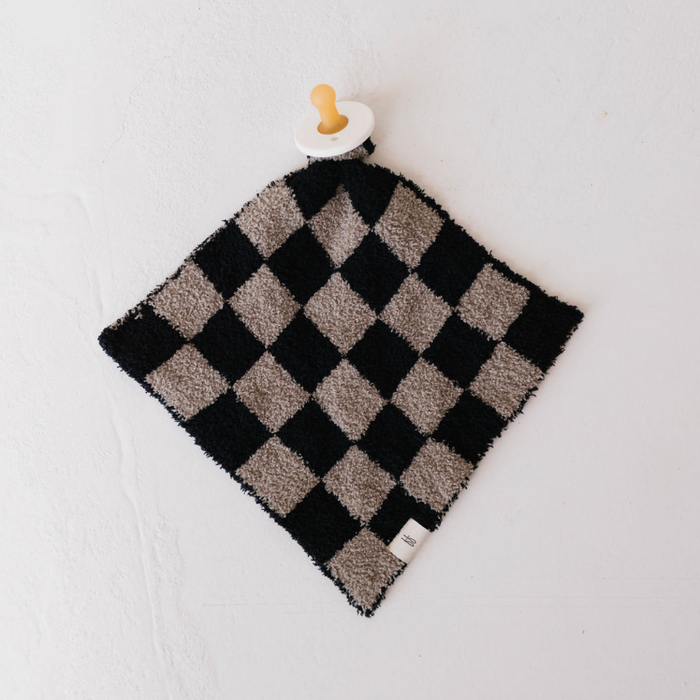 The "Lovey | Checkmate Checkerboard" by forever french baby, a luxurious microfiber feather yarn security blanket in a black and white checkerboard pattern, elegantly graces a light-colored surface. It features an attached wooden ring and pacifier at one corner—an exquisite creation for your little one.