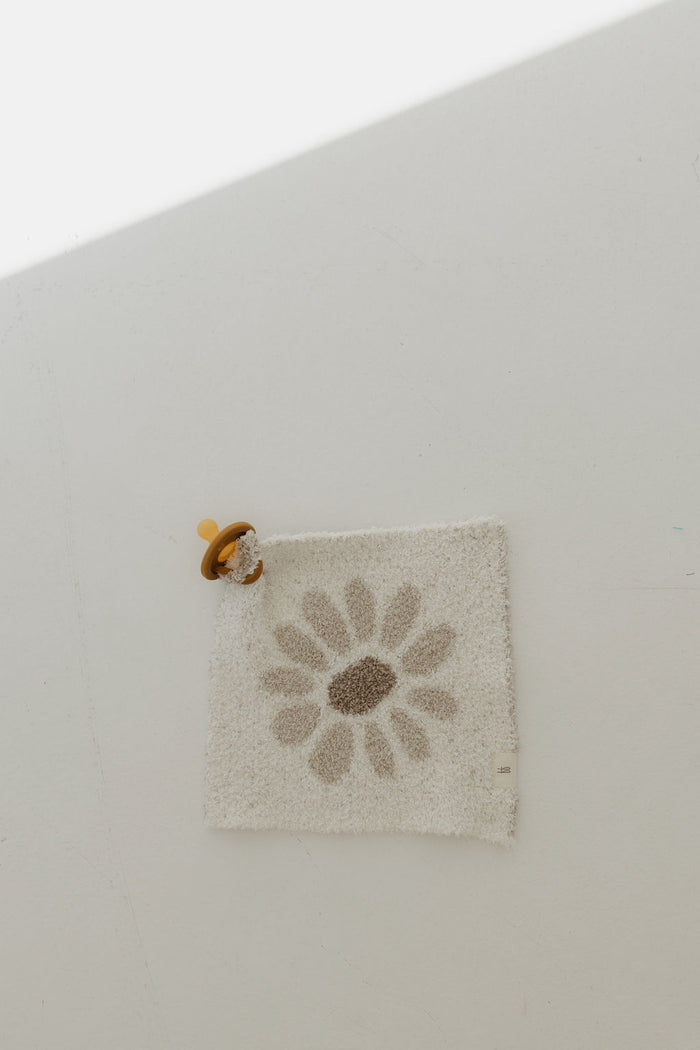 The Lovey | Daisy Fields by forever french baby lies on a light surface. A brown pacifier with a yellow ring is attached to one corner of the square white mat. Crafted from microfiber feather yarn, it glows as sunlight streams in at an angle.