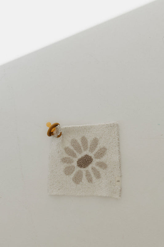 The Lovey | Daisy Fields by forever french baby lies on a light surface. A brown pacifier with a yellow ring is attached to one corner of the square white mat. Crafted from microfiber feather yarn, it glows as sunlight streams in at an angle.