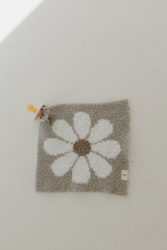 On a light surface lies the "Lovey | Darlin'" by forever french baby, a soft square gray baby blanket crafted from microfiber feather yarn, featuring a white daisy pattern with brown centers and a yellow pacifier attached to one corner.
