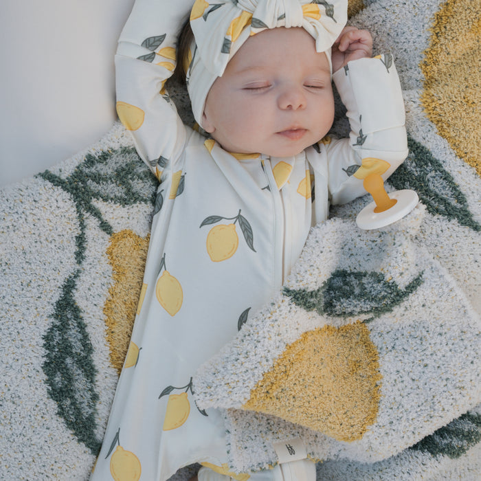 A baby sleeps on a blanket with yellow lemons, wearing a lemon-patterned forever french baby onesie and headband. Nearby are a pacifier and the *PRE-ORDER* Lovey | Lemon from forever french baby as the baby rests with arms raised.